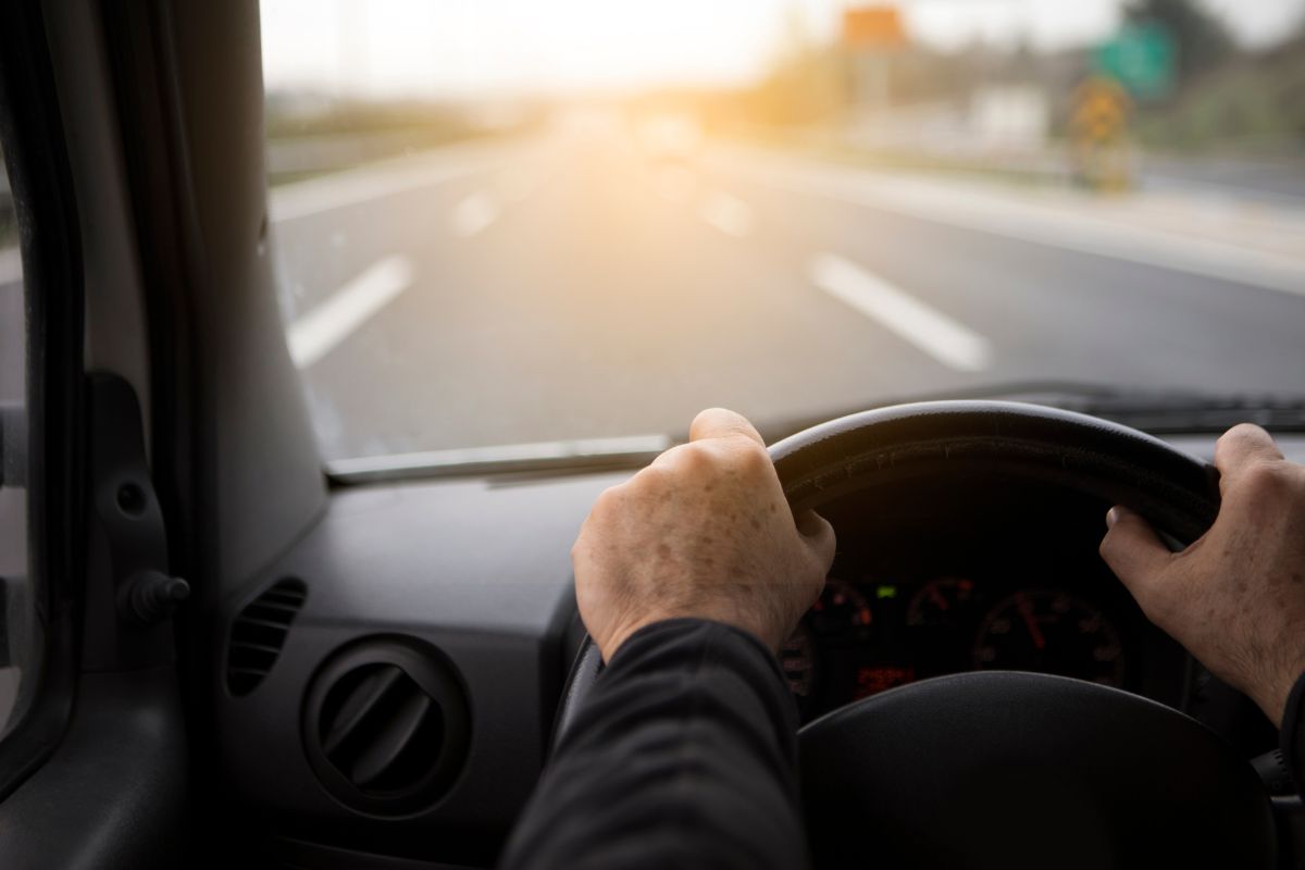 Mãos de um homem no volante de um carro dirigindo em uma estrada vazia