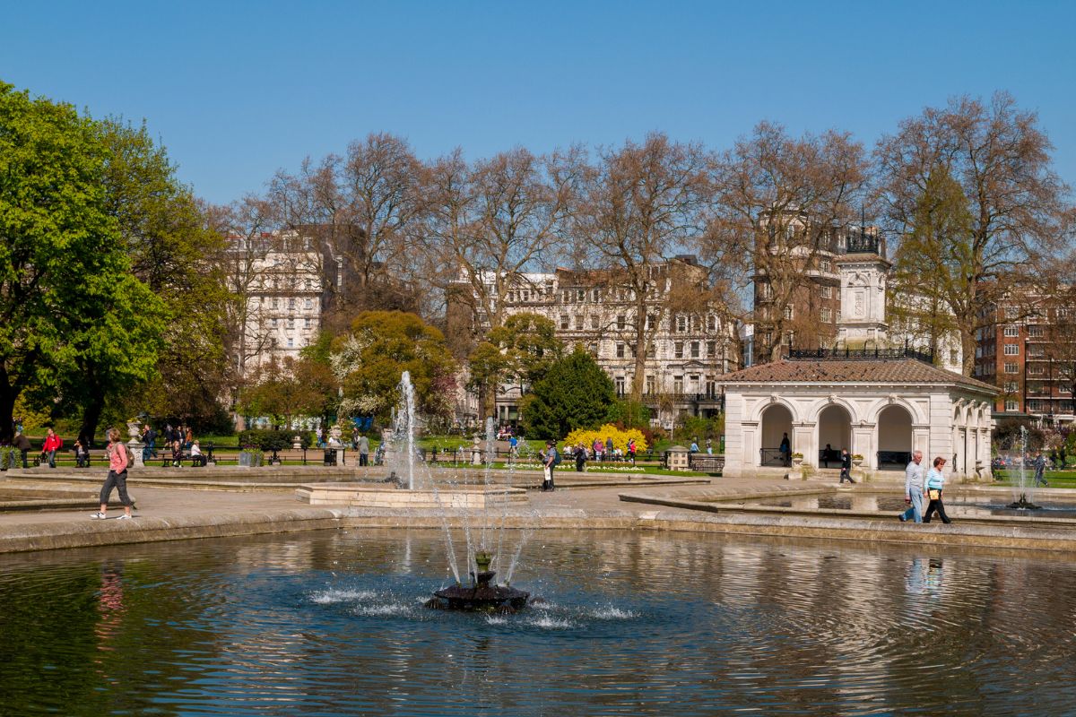 Hyde Park, de Londres, com parte do lago artificial, um chafariz, pessoas caminhando no entorno e edifícios da cidade ao fundo