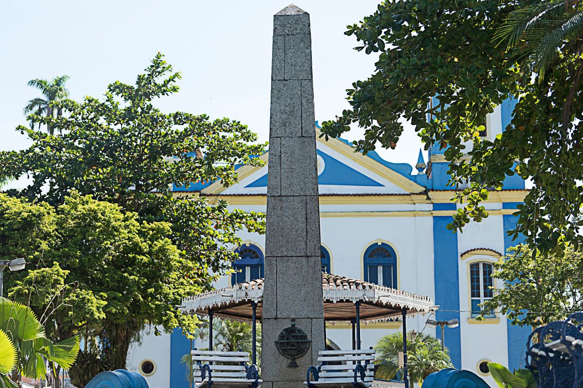Igreja no estilo colonial pintada de branco, amarelo e azul, com um pequeno coreto em frente e um monumento no formato de obelisco