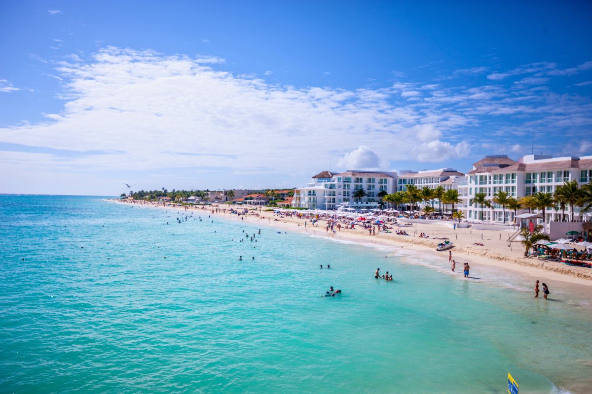 Vista aérea da Playa del Carmen com mar de água calma e transparente e banhistas tanto na areia quanto nadando, além de estrutura hoteleira ao fundo