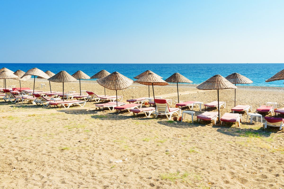 Praia de Çirali, na Turquia, com areia grossa, espreguiçadeiras e guarda-sóis de palha em frente ao mar de água tranquila e azul turquesa