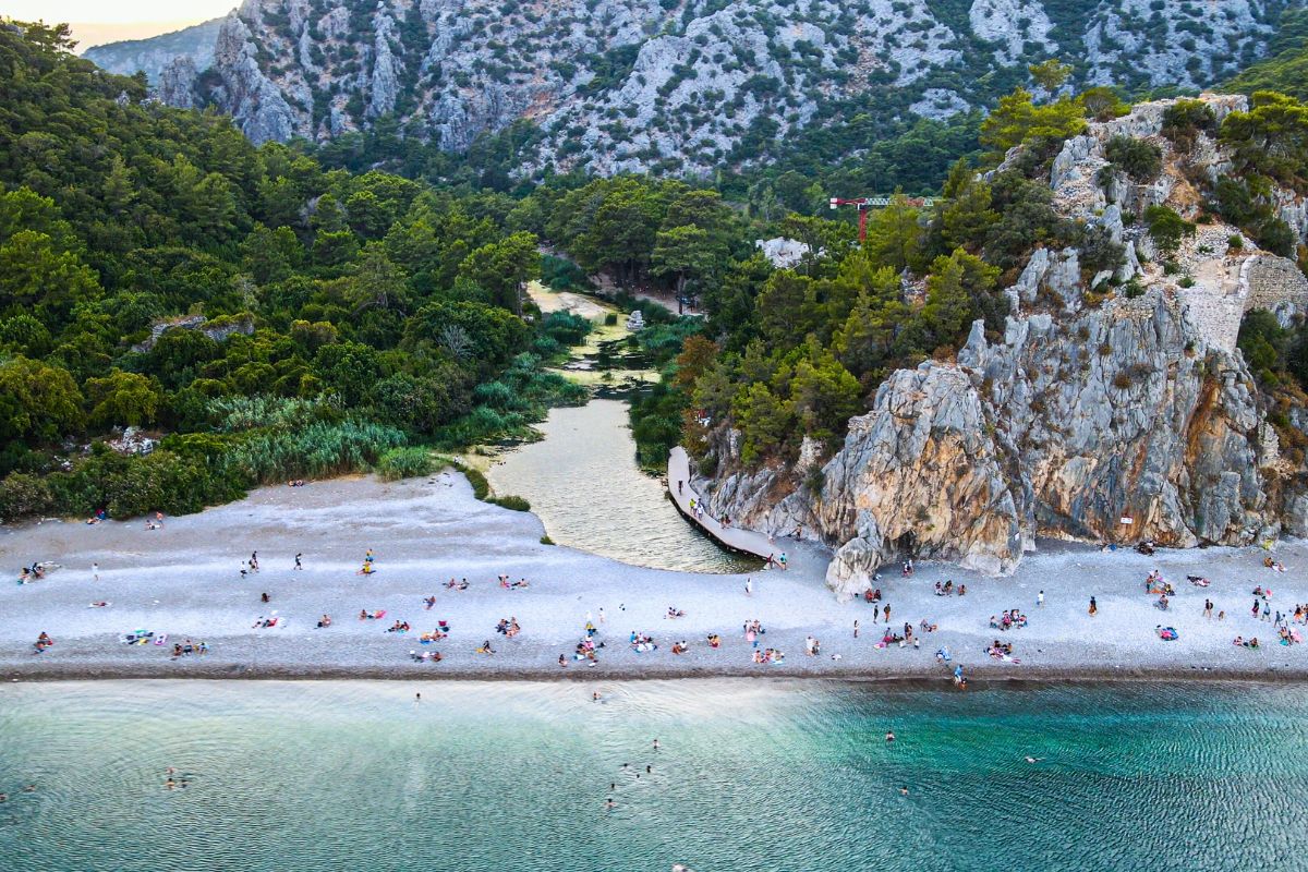 Praia de Olympos, na Turquia, vista de cima com o mar calmo em primeiro plano, a faixa de areia com poucos banhistas em segundo e, em terceiro plano, a floresta densa e o relevo montanhoso ao fundo