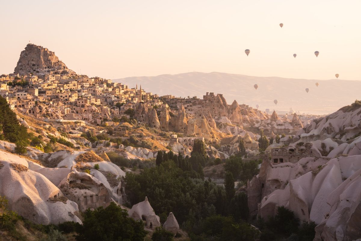 Pequena vila da Capadócia vista de longe com castelo de pedras no topo da colina e balões ao longe no horizonte